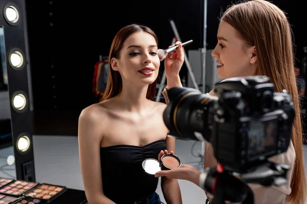 化妆师在数码相机附近的快乐模型上涂面部粉的选择焦点 — 图库照片