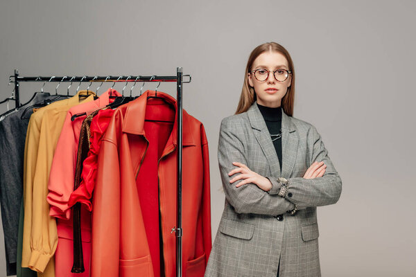 attractive stylist in glasses standing with crossed arms near trendy clothing isolated on grey 