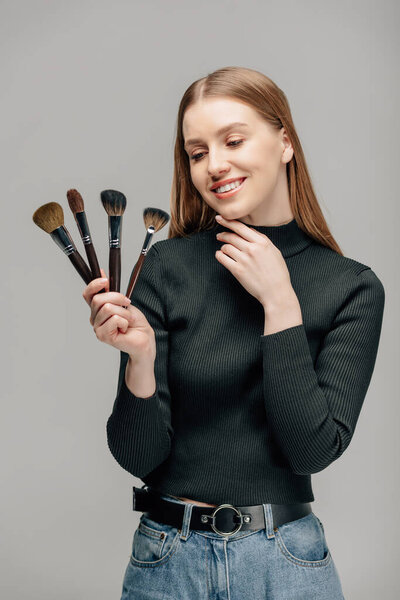 happy makeup artist holding cosmetic brushes isolated on grey 