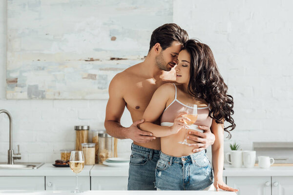 Man hugging girl with closed eyes and glass of wine from behind in kitchen