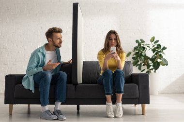 Kafası karışmış bir adam kız arkadaşına bakıyor. Kanepede akıllı telefonun büyük bir modelinin yanında sohbet ediyor. 