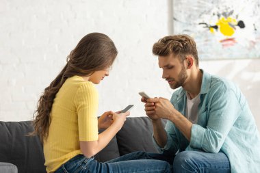 Evde kanepede otururken akıllı telefon kullanan genç çiftin yan görüntüsü 