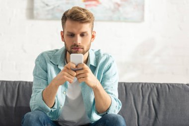 Evdeki kanepede otururken akıllı telefon kullanan yakışıklı adam. 