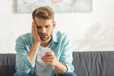 Oturma odasındaki koltukta akıllı telefon kullanan yorgun adam.