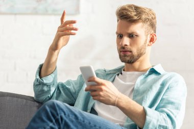 Kanepede akıllı telefon kullanırken kafası karışmış bir adam el kol hareketi yapıyor. 