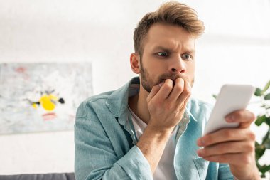Oturma odasında akıllı telefon kullanan eli ağıza yakın korkmuş bir adamın seçici odağı. 