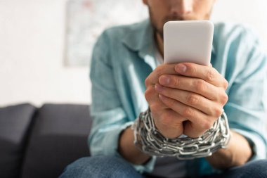 Selective focus of man with tied hands with chain using smartphone on couch  clipart