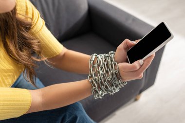 Eli kolu bağlı, demir zincirli, akıllı telefonu koltukta tutan kadın görüntüsü.