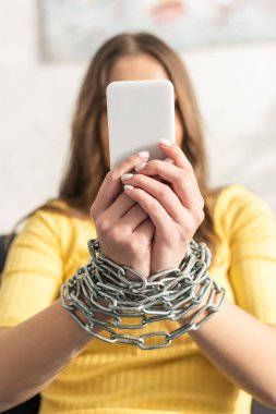 Metal zincirli, akıllı telefonu tutan elleri bağlı genç bir kadının seçici odağı. 
