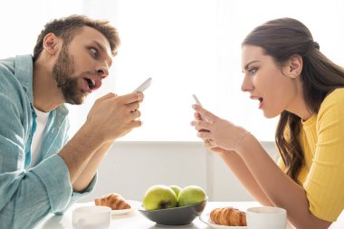 Kahvaltı masasının yanında akıllı telefon kullanan şok olmuş çiftin yan görüntüsü 