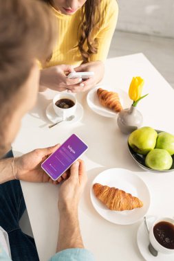 KYIV, UKRAINE - 21 Şubat 2020: Instagram uygulamalı akıllı telefon kullanan erkeğin seçici odak noktası mutfakta kahvaltı sırasında kız arkadaşının sohbet etmesi 