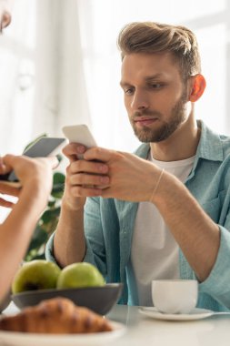 Kahve ve kruvasanın yanında akıllı telefon tutan bağımlı çiftin seçici odak noktası. 