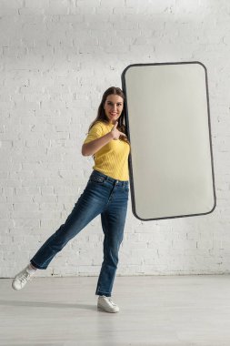 Gülümseyen kız beyaz tuğla duvarın yanındaki akıllı telefon modelini işaret ediyor.
