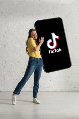 KYIV, UKRAINE - 21 Şubat 2020: Beyaz tuğla duvarın yanında TikTok uygulaması olan akıllı telefonun modelini tutan mutlu kadın