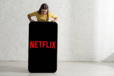 KYIV, UKRAINE - 21 Şubat 2020: Beyaz tuğla duvarın yanındaki akıllı telefon modelinde netflix uygulamasına bakan kadın