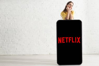 KYIV, UKRAINE - 21 Şubat 2020: Gülümseyen kız evinde netflix uygulaması olan akıllı telefonun büyük bir modelinin yanında kameraya bakıyor 