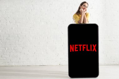 KYIV, UKRAINE - FEBRUARY 21, 2020: Smiling girl looking at camera near big model of smartphone with netflix app at home  clipart