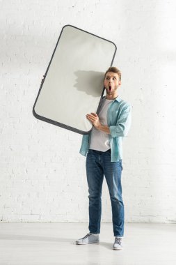 Shocked man holding model of smartphone near white brick wall  clipart