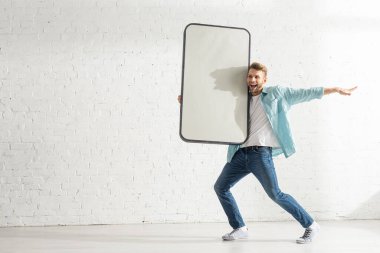 Beyaz tuğla duvarın yanında akıllı telefonun büyük bir modelini tutarken gülen neşeli adam.