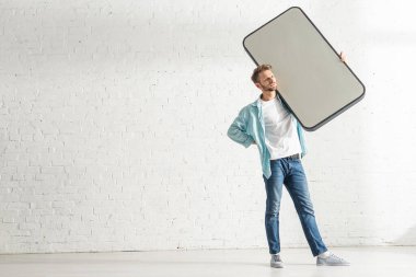 Beyaz tuğla duvarın yanında elinde büyük bir akıllı telefon modeli tutan yakışıklı bir adam.