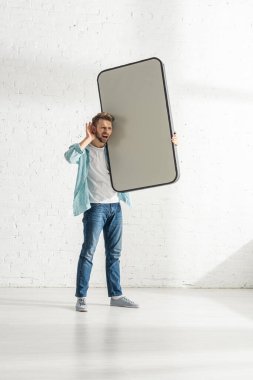 Eli kulağında yakışıklı bir adam beyaz tuğla duvarın yanında büyük bir akıllı telefon modeli tutuyor.