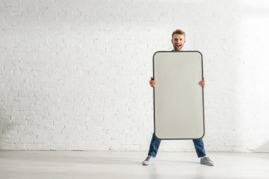 Neşeli adam beyaz tuğla duvarın yanında akıllı telefonun büyük bir modelini tutuyor. 
