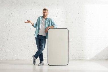 Gülümseyen adam akıllı telefonun büyük bir modelinin yanında omuz silkiyor. 