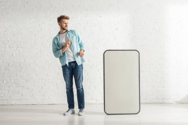 Beyaz tuğla duvarın yanında büyük bir akıllı telefon mankenine bakan korkmuş bir adam.