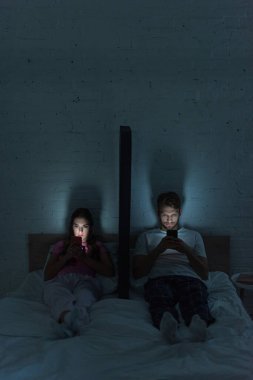 Akıllı telefonları olan genç bir çift gece yatağında akıllı telefon modelinin yanında oturuyor. 