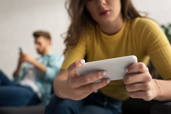 Selective focus of girl with smartphone sitting near boyfriend on couch 