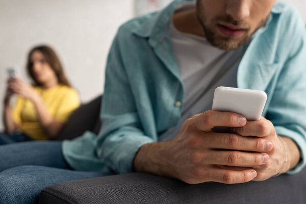 Selective focus of man using smartphone near girlfriend on couch 