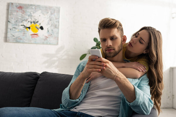 Beautiful woman embracing addicted boyfriend with smartphone on sofa 