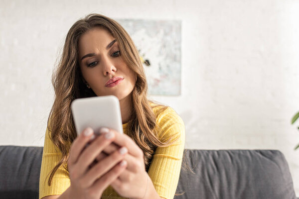 Selective focus of confused girl using smartphone on couch in living room