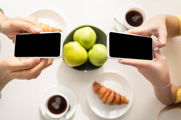 Вид сверху пары, использующей смартфоны рядом с завтраком на столе
 