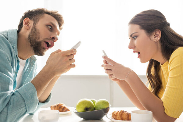 Side view of shocked couple using smartphones near breakfast on table 