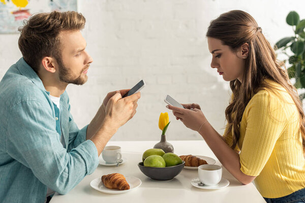 Side view of man chatting near girlfriend holding smartphone during breakfast in kitchen 