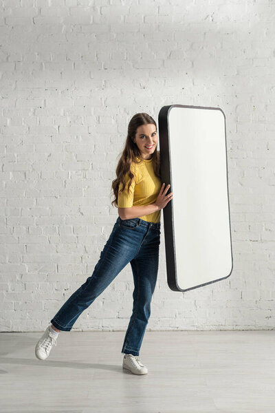 Beautiful girl smiling at camera while holding model of smartphone at home 