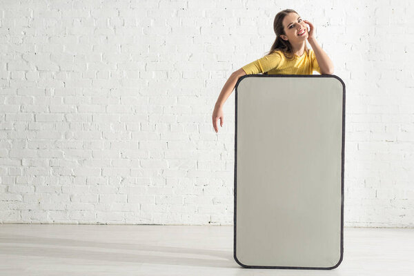 Красивая женщина с рукой возле щеки улыбается в камеру рядом с большой моделью смартфона
 