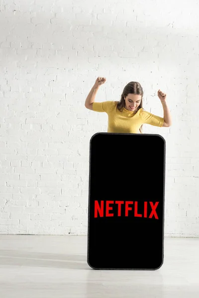 stock image KYIV, UKRAINE - FEBRUARY 21, 2020: Smiling girl showing yes gesture and looking at model of smartphone with netflix app