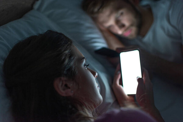 Selective focus of smartphone dependent girl lying near boyfriend on bed at night 
