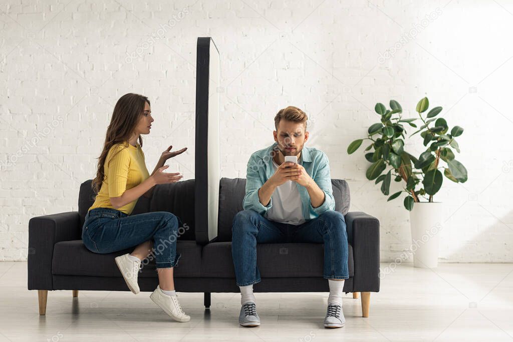 Confused girl looking at big model of smartphone near addicted boyfriend chatting on sofa 