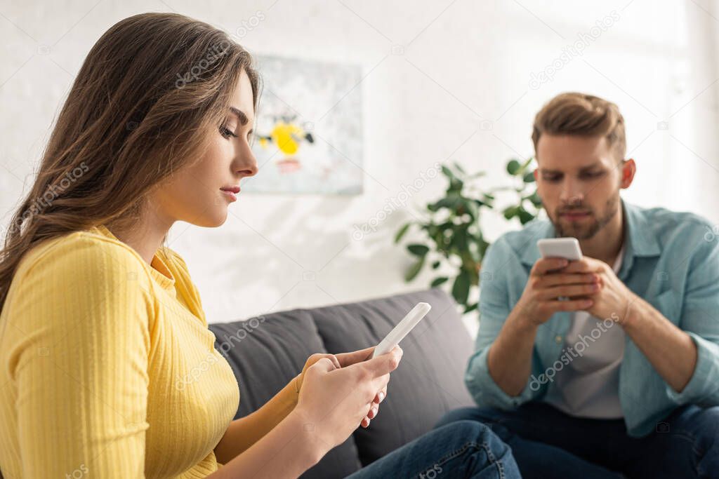 Selective focus of beautiful woman using smartphone near man on couch at home 