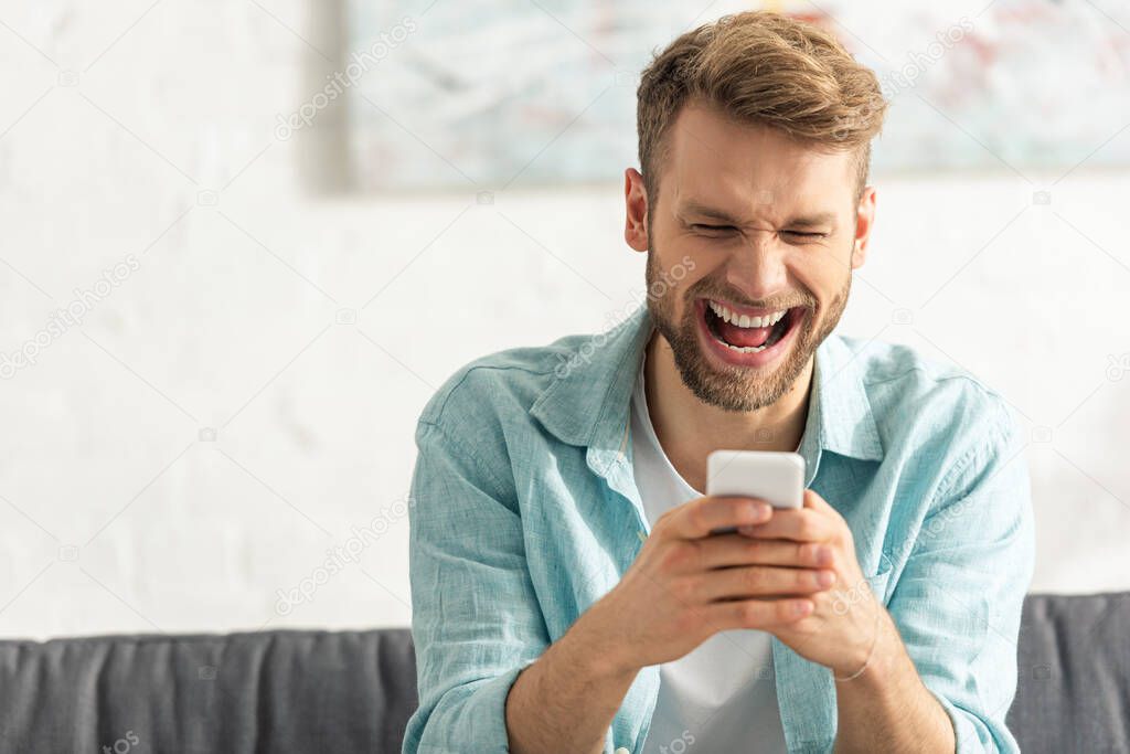 Selective focus of cheerful man laughing while using smartphone on couch 