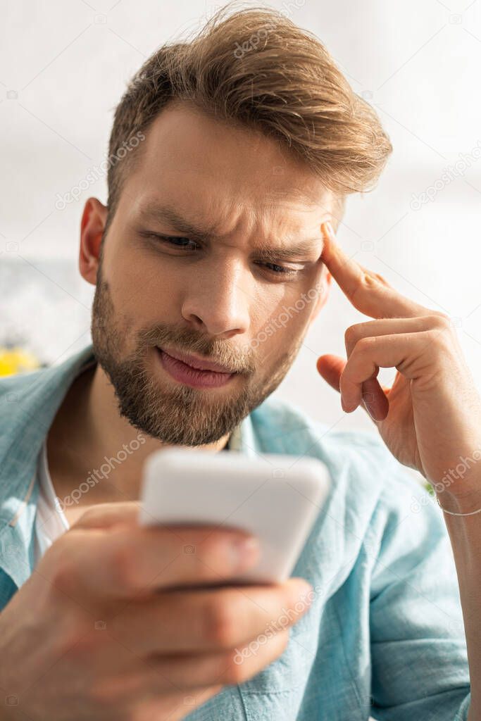 Selective focus of thoughtful man with fingers near forehead using smartphone 