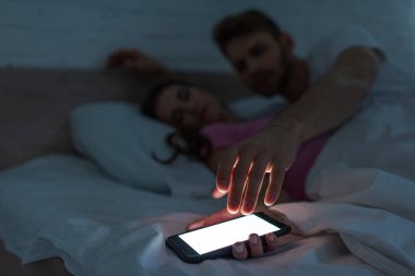 Seçici odak noktası, gece yatakta erkek arkadaşının yanında akıllı telefon tutan kadın. 