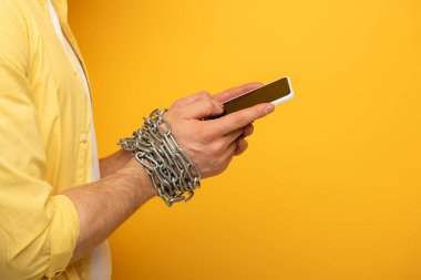 Cropped view of man with metal chain around hands holding smartphone with blank screen on yellow background clipart