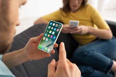 KYIV, UKRAINE - 21 Şubat 2020: Akıllı telefonu elinde tutan erkeğin seçici odak noktası iPhone ekranı ile kadının yanındaki kanepe 