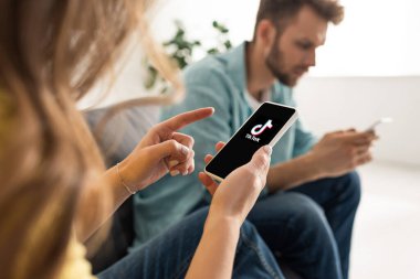 KYIV, UKRAINE - FEBRUARY 21, 2020: Selective focus of woman pointing with finger at smartphone with TikTok app near boyfriend chatting on couch  clipart
