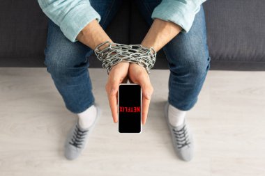 KYIV, UKRAINE - 21 Şubat 2020: Kanepede netflix uygulamalı akıllı telefonu elinde metal zincir tutan adamın üst görünümü 