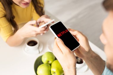 KYIV, UKRAINE - 21 Şubat 2020: Akıllı telefonu kız arkadaşının yanında netflix uygulaması ve masada kahveyle tutan adamın seçici odak noktası 